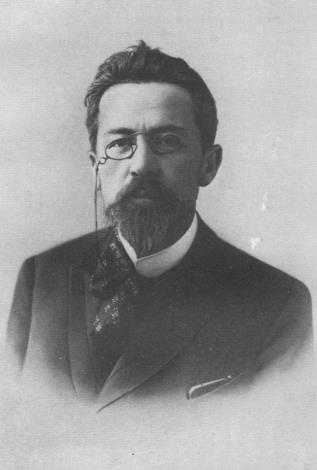 Man with dark hair and beard, wearing round spectacles and a monocle. He is dressed in a dark suit with a high-collared shirt and a patterned tie. The photo is black and white and has a formal, early 20th-century style.